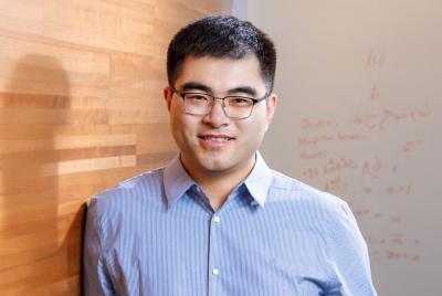 Yizhong Wang in a blue botton-down shirt leaning against a wood slat wall, smiling at the viewer.