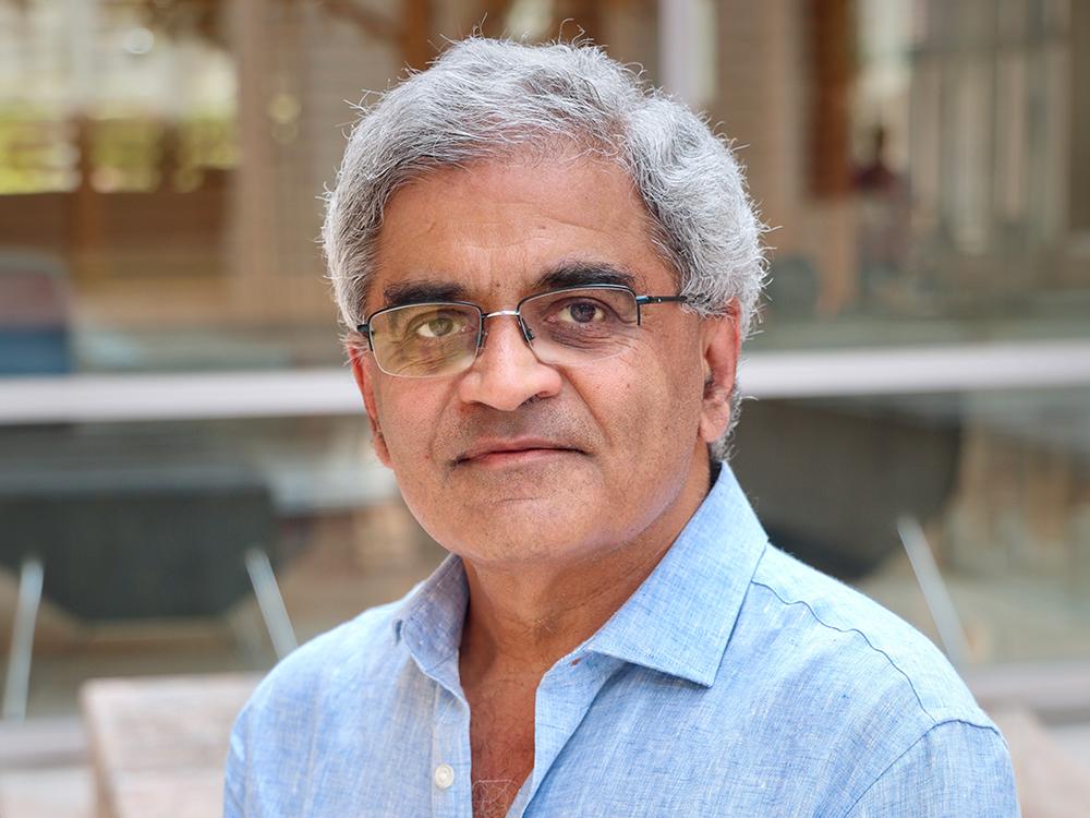 Professor Keshav Pingali in front of the Gates Dell Complex in a light blue denim button-down shirt and black framed glasses.