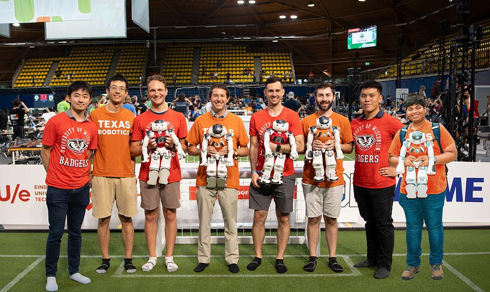 Eight people, half of them in Texas Robotics shirts and half in Wisconsin badgers t-shirts, pose on a soccer field, several of them holding small robots in jerseys.