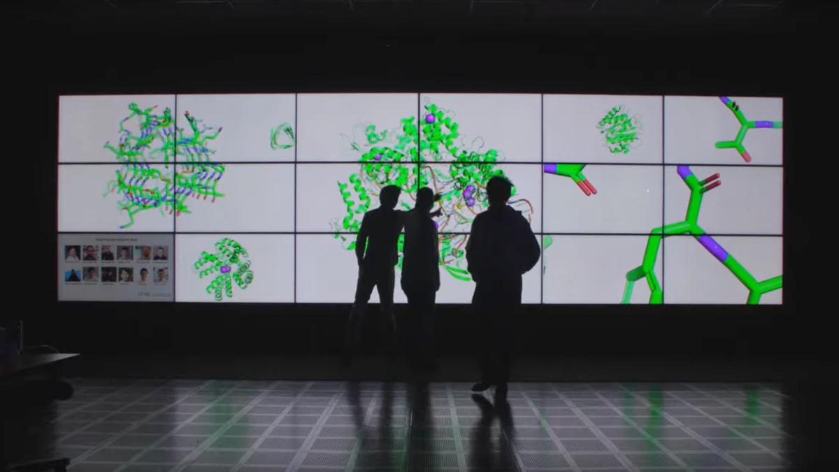 Three people stand silhouetted  in front of a wall-sized video display that shows several large colorful illustrations of molecules