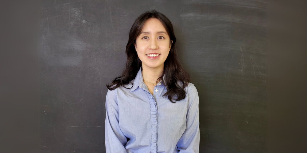 Mina Huh standing against a blackboard wearing a blue button-down shirt.