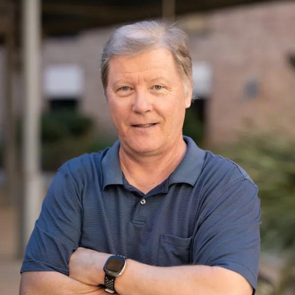 Risto Miikkulainen standing in front of a UT building in a blue polo style shirt with arms crossed.