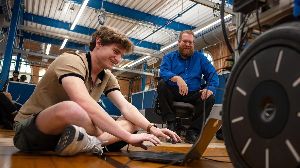 A student sits on the floor of a large laboratory, typing on a laptop while a professor sits nearby overseeing his work