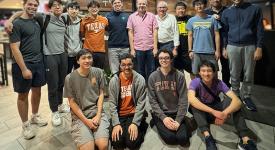 The UT Programming Team gathered together for a photo in a restuarant. Eight students and three faculty coaches stand in the back row. Four students sit in the front row.