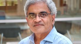 Professor Keshav Pingali in front of the Gates Dell Complex in a light blue denim button-down shirt and black framed glasses.