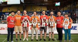 Eight people, half of them in Texas Robotics shirts and half in Wisconsin badgers t-shirts, pose on a soccer field, several of them holding small robots in jerseys.