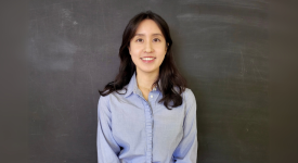 Mina Huh standing against a blackboard wearing a blue button-down shirt.