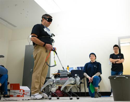 A robotic guide dog being tested by a man standing with sensors on his arms and blindfold over his eyes.