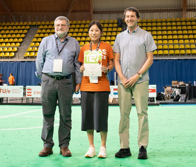 A woman in a Texas Robotics shirt holds up a certificate while posing between two men on an indoor soccer field.