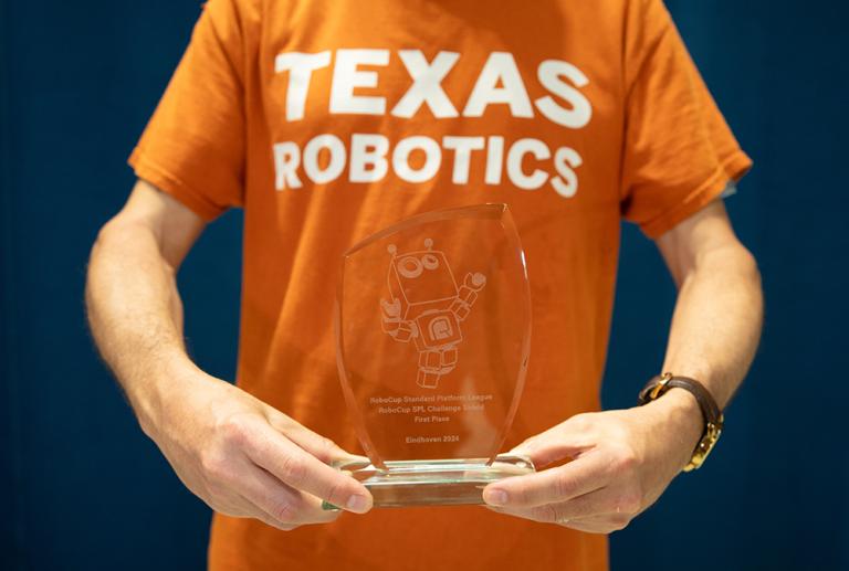 A person in a Texas Robotics t-shirt holds a trophy that reads &quot;RoboCup Standard Platform League Challenge Shield 1st Place: Eindhoven 2024&quot; 