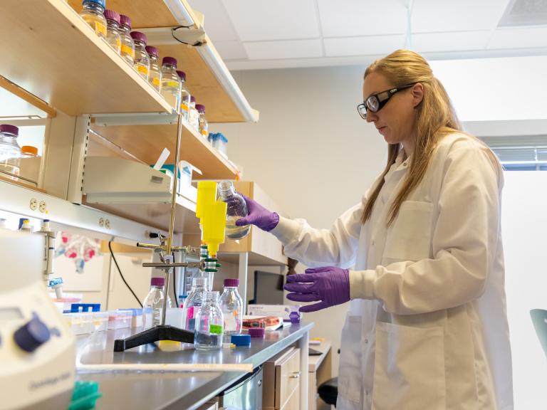 A scientist in a white lab coat stands at a lab bench and pours a clear liquid into a glass jar